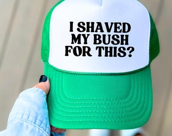 Può includere: Un cappello da camionista verde e bianco con la scritta "I shaved my bush for this?" in nero. Il cappello ha un pannello frontale bianco e un retro in rete verde. La visiera e il bottone superiore sono anch'essi verdi.