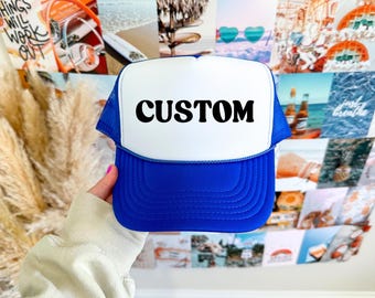 May include: A blue and white trucker hat with the word "CUSTOM" in black block letters. The hat has a blue brim and mesh back, and a white front panel. The hat is held by a person.