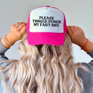 Peut inclure: Casquette de camionneur rose et blanche avec le texte "PLEASE TONGUE PUNCH MY FART BOX" en noir. La casquette a un dos en filet et une visière incurvée. La casquette est portée par une personne aux cheveux blonds.