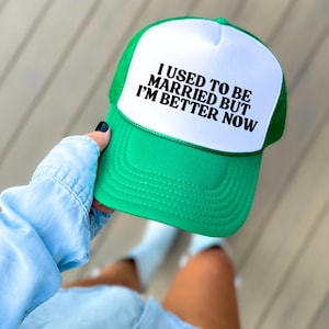 May include: A green and white trucker hat with the text "I USED TO BE MARRIED BUT I'M BETTER NOW" in black. The hat has a curved brim and a mesh back.