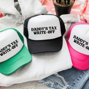 May include: Three trucker hats with the text "DADDY'S TAX WRITE-OFF" in bold black letters. The hats have white fronts and black, green, and pink brims. The hats are on top of a grey hoodie and denim.