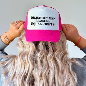 May include: A pink and white trucker hat with the text "OBJECTIFY MEN BECAUSE EQUAL RIGHTS" in black. The hat has a mesh back and a curved brim. The hat is being held by a person with blonde hair.