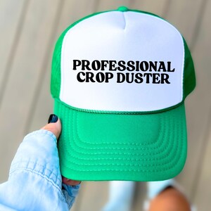 May include: A green and white trucker hat with the text "PROFESSIONAL CROP DUSTER" in bold black letters. The hat has a green mesh back and a white front panel. The brim is green.