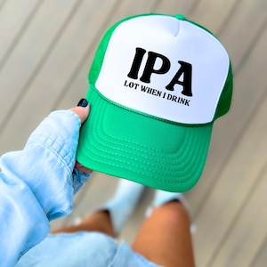 May include: A green and white trucker hat with the text "IPA LOT WHEN I DRINK" in black. The hat has a green brim and mesh back, with a white front panel. The text is in a bold, sans-serif font.