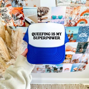 May include: A blue and white trucker hat with the text "QUEEFING IS MY SUPERPOWER" in black. The hat has a mesh back and a curved brim. The hat is held in front of a collage of photos.
