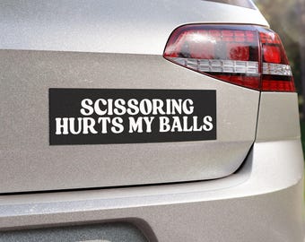 Può includere: Un adesivo rettangolare nero per paraurti con testo bianco che recita SCISSORING HURTS MY BALLS in un carattere grassetto senza grazie. L'adesivo è applicato sul bagagliaio argentato di un veicolo, sotto il lunotto posteriore.