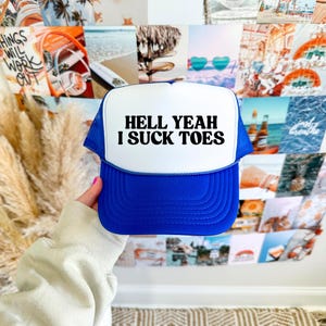 Könnte beinhalten: Blau-weiße Trucker-Mütze mit dem Text "HELL YEAH I SUCK TOES" in Schwarz. Die Mütze hat einen Mesh-Rücken und einen gebogenen Schirm. Die Mütze wird vor einer Collage von Fotos gehalten.