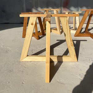 May include: A set of light-colored wooden table bases featuring a trapezoidal frame design with a central vertical support beam. The structures are constructed from natural pine wood with visible grain patterns, arranged in a group on a flat surface.