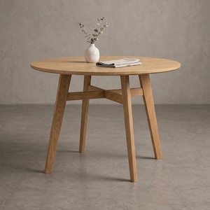 Peut inclure: Une table à manger ronde en bois clair dotée de quatre pieds fuselés et d'une structure de support en croix. Un petit vase en céramique blanche contenant un brin d'eucalyptus séché et une serviette en tissu pliée reposent sur le plateau.