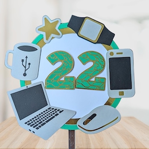 May include: A birthday cake topper with a white background and a gold and green border. The number "22" is in the center, surrounded by cutouts of a coffee mug, star, smartwatch, smartphone, laptop, and mouse. The number "22" has a circuit board design.