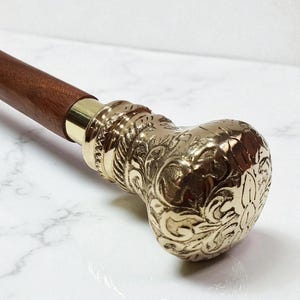 Puede incluir: Un bastón de madera con un mango de latón. El mango está intrincadamente diseñado con patrones florales. El eje es de color marrón oscuro y el mango de latón tiene un acabado dorado. Un accesorio clásico.