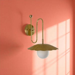 May include: A brass wall sconce with a unique design. It features a round base, a curved arm, and a scalloped shade. The shade holds a white, spherical light. The sconce is mounted on a coral-colored wall.