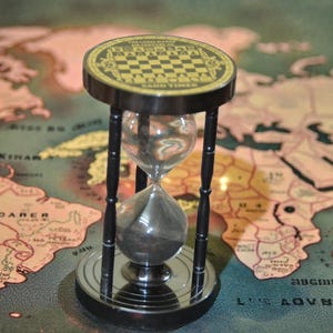 May include: An hourglass with a black frame and a checkerboard design on top. The glass bulb contains dark sand. The hourglass is set against a vintage world map background.