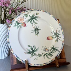 Może przedstawiać: Biały ceramiczny talerz z kolorową obwódką i botanicznym wzorem z owocami męczennicy i zielonymi liśćmi. Talerz jest prezentowany na drewnianym stojaku, z wazonem kwiatów w tle.