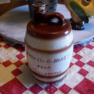 May include: A brown and white ceramic honey jar with a cork stopper. The jar is shaped like a barrel and has the text "Barrel-O-Honey From The Ozarks" written on it.