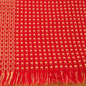 May include: A woven textile blanket featuring a vibrant red background with a repeating cream-colored geometric pattern. The left section displays a grid of four-pointed shapes, while the right side shows small vertical dashes. The edges have fringe.