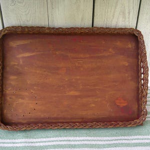 May include: Rectangular wooden serving tray with a woven border. The tray has a dark brown painted surface with visible brushstrokes and a braided edge. The tray is sitting on a striped green and white fabric.
