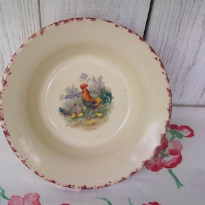 May include: A white ceramic bowl with a red rim and a painted design of a rooster and chicks. The bowl is on a white and red tablecloth.