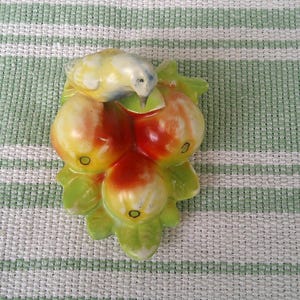 May include: A ceramic figurine of a bird perched on a cluster of apples. The bird is painted in shades of yellow and blue, while the apples are red and yellow with green leaves. The figurine is on a green and white striped background.