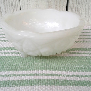 May include: A white, vintage milk glass bowl with a scalloped rim and three small feet. The bowl has a textured, floral design around the base. It sits on a green and white striped fabric, with a white wooden background.