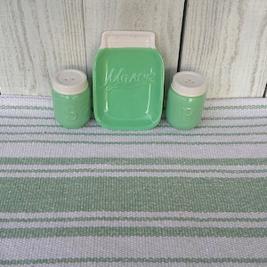 May include: A mint green ceramic salt and pepper shaker set with a matching jar-shaped tray. The tray is inscribed with the word "Mason". The shakers and tray have white lids. The set is displayed on a green and white striped mat.