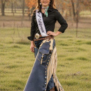 May include: A woman wearing a black long-sleeve shirt, blue jeans, and a white cowboy hat. She is wearing a sash that says "Miss Rodeo California". She is also wearing blue chaps with fringe.