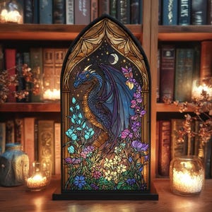 May include: Stained glass panel depicting a blue and purple dragon surrounded by flowers and foliage. The dragon is set against a starry night sky with a crescent moon. The panel is framed in black and sits on a black base.