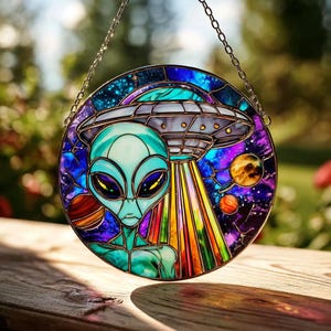May include: Round stained glass window featuring an alien with a teal face and body, large black eyes, and a gray spaceship. The background is a mix of blue, purple, and orange, with planets and stars. A silver chain is attached for hanging.
