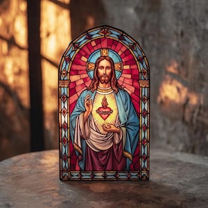 Puede incluir: Un panel de vidrieras que representa una figura religiosa con un corazón brillante. La obra de arte presenta una vibrante paleta de colores rojo, azul y dorado, con un marco oscuro. El panel está iluminado, creando un brillo cálido y acogedor.