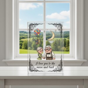 Può includere: Blocco in acrilico trasparente con un'illustrazione a cartoni animati di una coppia di anziani che si tengono per mano, una casa con palloncini, una luna crescente e la scritta "I love you to the moon and back". Bordo floreale decorativo.