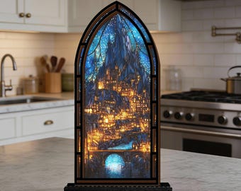 Può includere: Lampada decorativa a forma di vetrata raffigurante una città fantasy notturna. La lampada ha una cornice in legno scuro e una base nera. La città è illuminata da luci gialle calde, su uno sfondo di cielo stellato blu. Un ponte attraversa un fiume.
