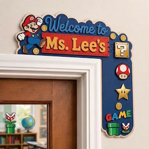 May include: A decorative L-shaped wooden door corner sign featuring Mario, a red banner with the text Welcome to Ms. Lee's, a yellow question block, a red and white mushroom, a yellow star, a green pipe, and the word GAME in colourful letters.