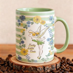 Op de afbeelding: Een keramische mok met een groene handgreep en rand, met een cartoon pixie-ontwerp. De mok heeft een witte achtergrond met groene strepen en bloemaccenten. De tekst "Pretty Little Pixie" is ook zichtbaar.