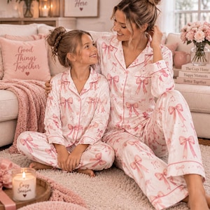 Op de afbeelding: Bijpassende tweedelige pyjamasets met een witte basis en een herhalend roze strikpatroon. Elke set bevat een overhemd met lange mouwen, knoopsluiting, een reverskraag en een borstzak, gecombineerd met een bijpassende lange broek met roze biesafwerking.
