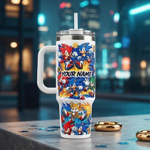 May include: A white stainless steel tumbler with a handle and a straw, featuring a colourful graphic print of Sonic the Hedgehog characters including Sonic, Tails, and Knuckles. The text "YOUR NAME" is printed in bold blue letters on the front.