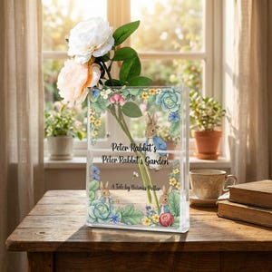 May include: A clear acrylic vase featuring illustrations of Peter Rabbit and floral designs. The vase holds white and peach-colored roses. Text on the vase reads "Peter Rabbit's Garden" and "A Tale by Beatrix Potter."