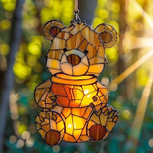 Puede incluir: Atrapasoles de oso de vidriera en tonos ámbar, marrón y amarillo. El oso sostiene un tarro de miel. El atrapasoles cuelga de una cadena dorada. El diseño del oso está hecho de muchos trozos pequeños de vidrio.