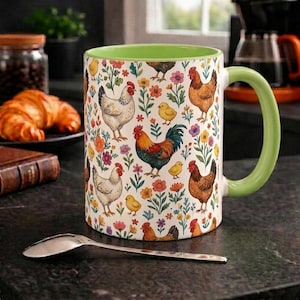 Puede incluir: Una taza de café de cerámica con asa y borde verdes. La taza presenta un patrón repetido de gallinas, gallos, pollitos y flores coloridas sobre un fondo blanco. Una cuchara plateada descansa sobre la encimera.