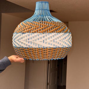 Puede incluir: Una lámpara colgante tejida con una combinación de colores azul, blanco y marrón. La lámpara está sujeta por la mano de una persona. La parte superior de la lámpara es azul, con bandas marrones y blancas. La lámpara está diseñada para colgar del techo.