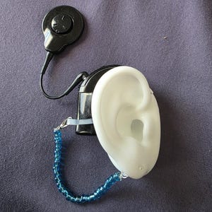 May include: A black cochlear implant processor with a round control panel is connected to a white ear mould. A blue beaded safety wire is attached to the ear mould. The items are on a purple fabric surface.