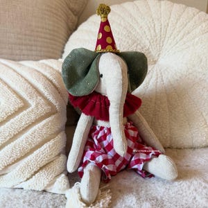May include: A stuffed elephant toy made of beige fabric, wearing a red and gold polka dot party hat, a dark red ruffled collar, and red and white gingham bloomers. The elephant has dark green corduroy ears and black bead eyes.