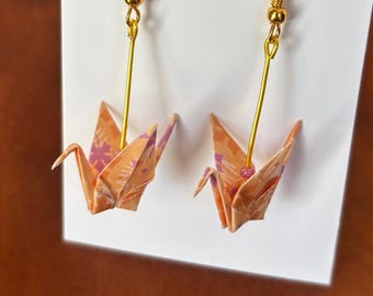 May include: Origami crane earrings with gold-colored hooks and wires. The cranes are made of orange paper with a floral pattern in pink and purple. The earrings are displayed on a white card.