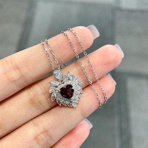 May include: A silver necklace featuring a heart-shaped pendant with a deep red center stone. The pendant is surrounded by clear, sparkling accent stones. The chain is delicate and silver, creating an elegant jewelry piece.