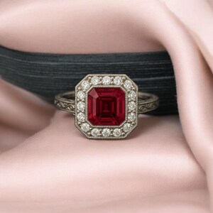 May include: A silver ring featuring a large, square, deep red gemstone, encircled by a halo of small, round, white gemstones. The ring's band has an ornate, engraved design. The ring is set against a soft pink background.