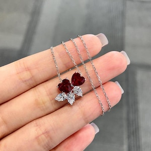 May include: A silver necklace featuring a cluster of heart-shaped, deep red gemstones and clear, pear-shaped stones. The pendant is suspended from a delicate chain. The necklace is displayed against a neutral background.