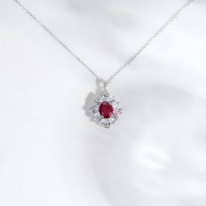 May include: A silver necklace with a pendant featuring a large, oval, deep red gemstone surrounded by smaller, clear gemstones. The pendant is suspended from a delicate silver chain, creating an elegant and eye-catching piece of jewelry.