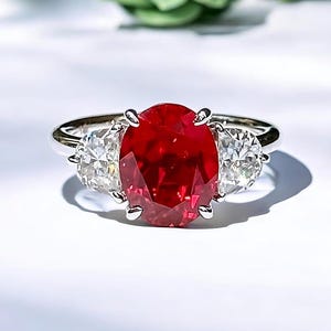 Könnte beinhalten: Ein silberner Ring mit einem großen, oval geschliffenen, leuchtend roten Edelstein und zwei kleineren, runden, klaren Edelsteinen. Der Ring ist vor einem hellen, weißen Hintergrund platziert, mit grünem Laub im Hintergrund.