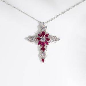 May include: A silver pendant necklace featuring a cross design. The cross is adorned with red gemstones and a central clear gemstone. The necklace hangs on a delicate silver chain, set against a white background.