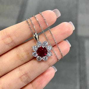 May include: A silver necklace featuring a flower-shaped pendant. The pendant has a large, round, deep red gemstone at its center, surrounded by smaller, clear gemstones. The chain is delicate and the pendant is held up by a small silver bail.
