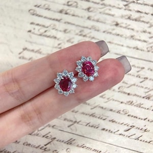 Könnte beinhalten: Ein Paar ovale Ohrringe mit Rubinen und Diamanten. Jeder Ohrring hat einen zentralen roten Rubin, umgeben von einem Kranz aus kleinen, funkelnden Diamanten. Die Ohrringe sind in einem silberfarbenen Metall gefasst.
